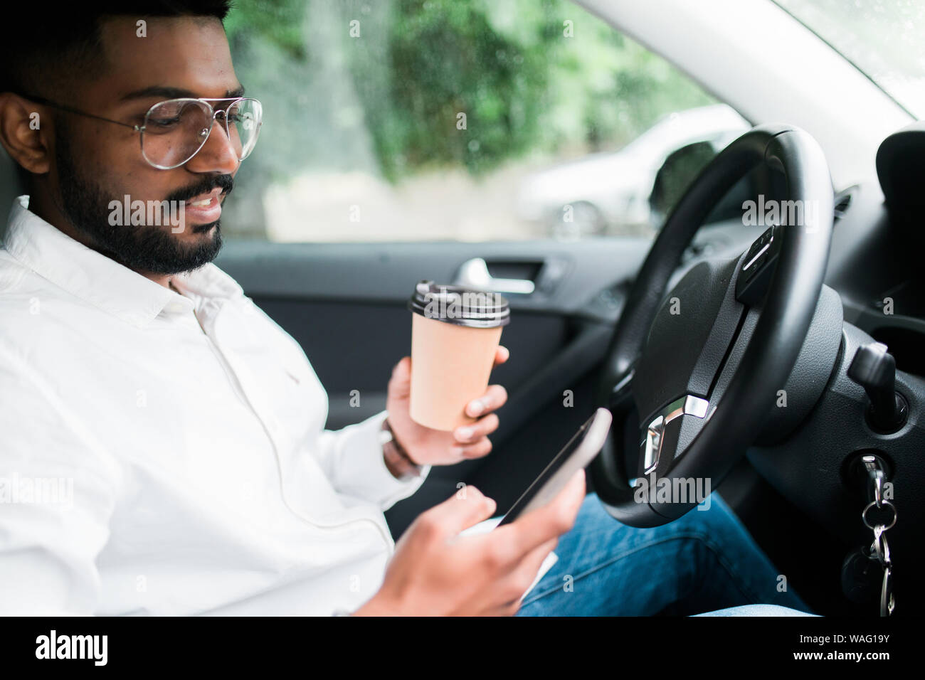 Stylish driver with a smartphone in hand and paper cup of hot coffee in the driver's seat. The concept of inattention at the wheel, rest, coffee break Stock Photo