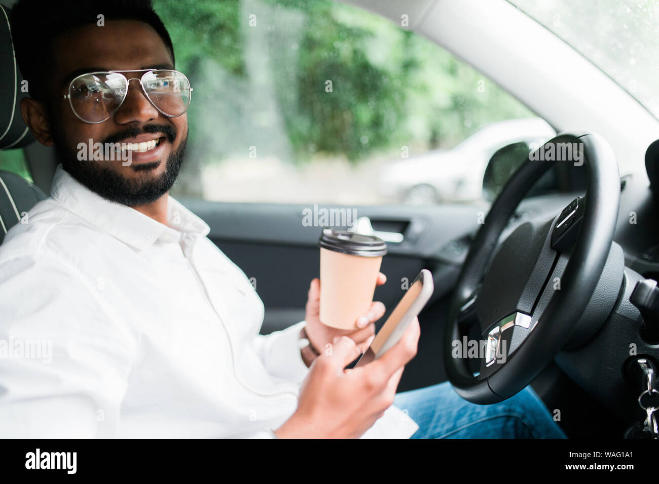 Stylish driver with a smartphone in hand and paper cup of hot coffee in the driver's seat. The concept of inattention at the wheel, rest, coffee break Stock Photo