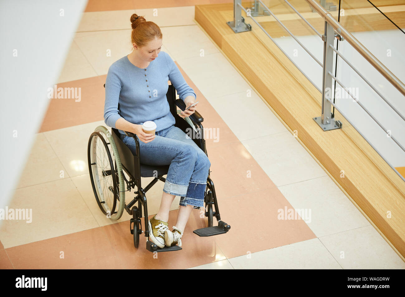 Young beautiful woman in wheelchair typing a message on mobile phone and drinking coffee while spending time in shopping centre Stock Photo