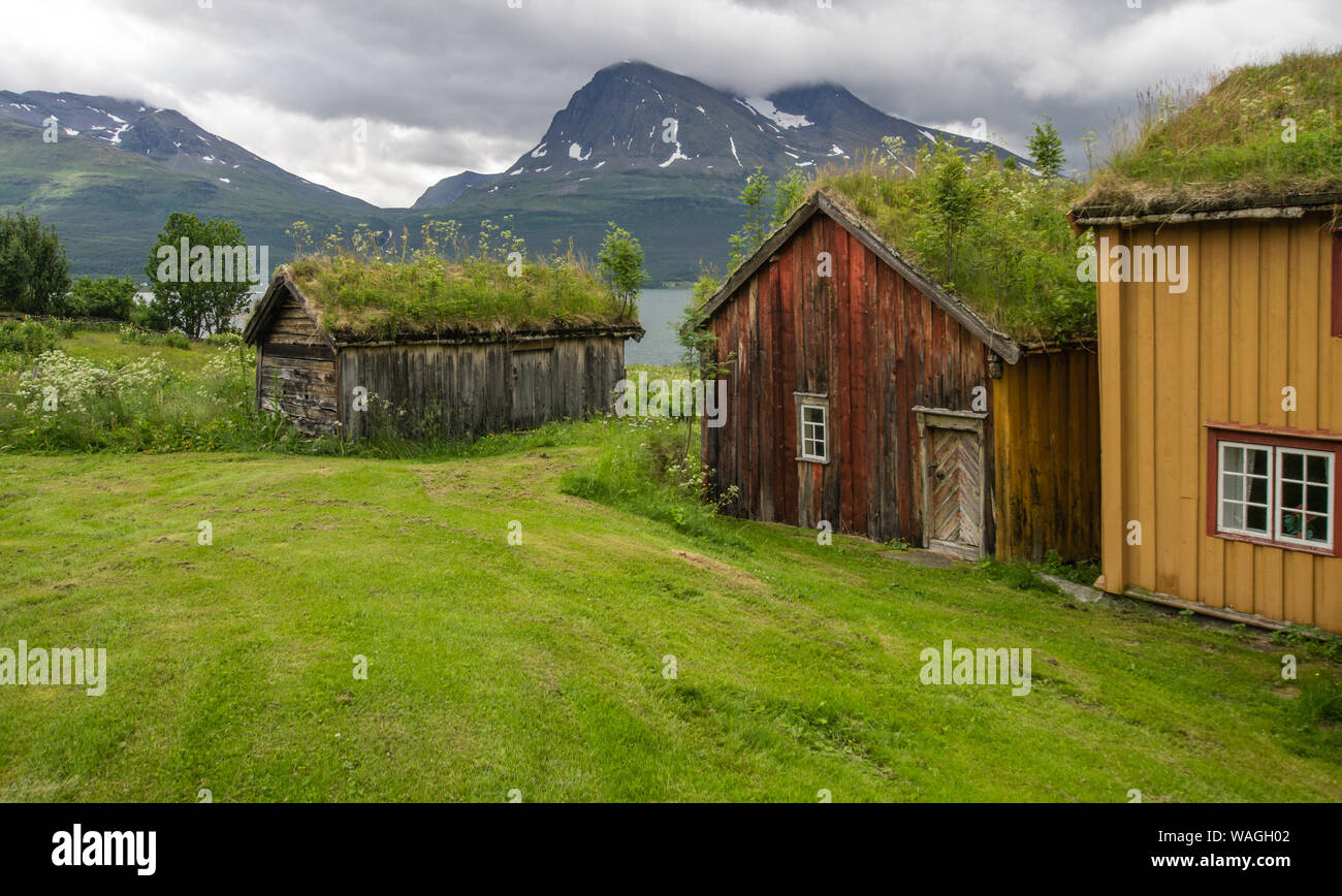 Historic wooden buildings in rural far north Norway - the roofs are overgrown by green vegetation, set at the shore of a fjord, mountains in backgroun Stock Photo
