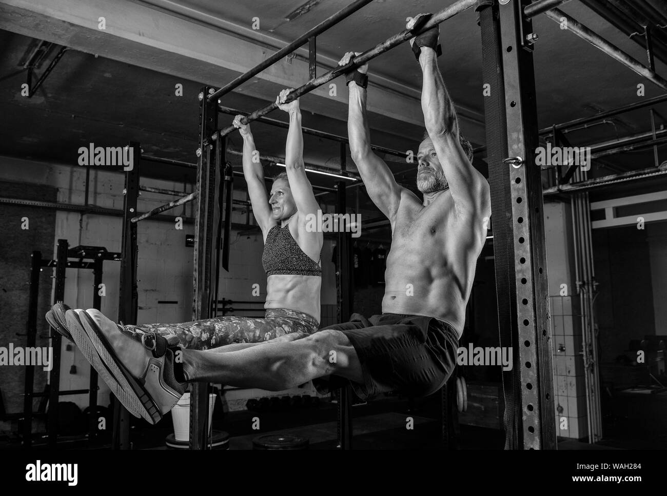 An older couple is doing a fitness training together and having fun. Muscular man and woman are doing the exercise l sit on the horizontal bar. Stock Photo