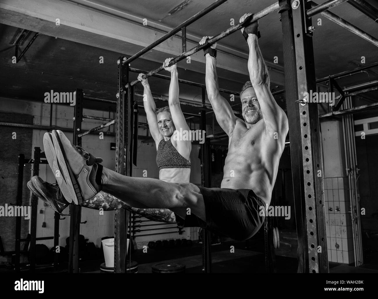 A middle aged couple is doing a fitness training together and having fun. Muscular man and woman are doing the exercise l sit on the horizontal bar. Stock Photo