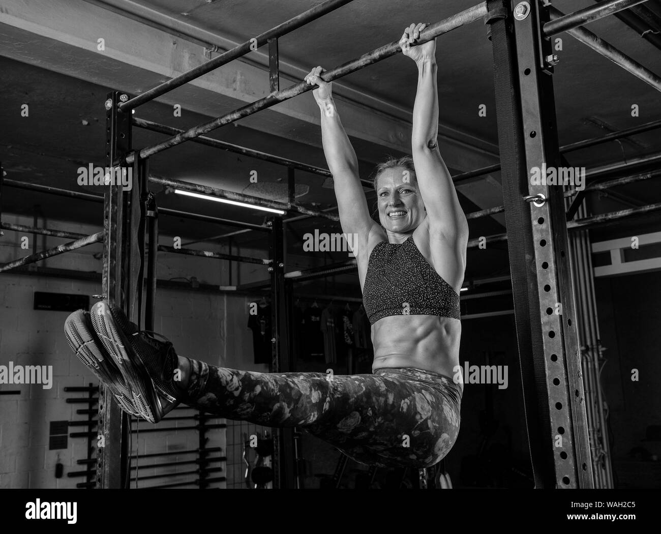 A beautiful middle aged blonde woman with strong abs is doing the exercise l sit on the horizontal bar. Functional fitness workout in a gym. Stock Photo