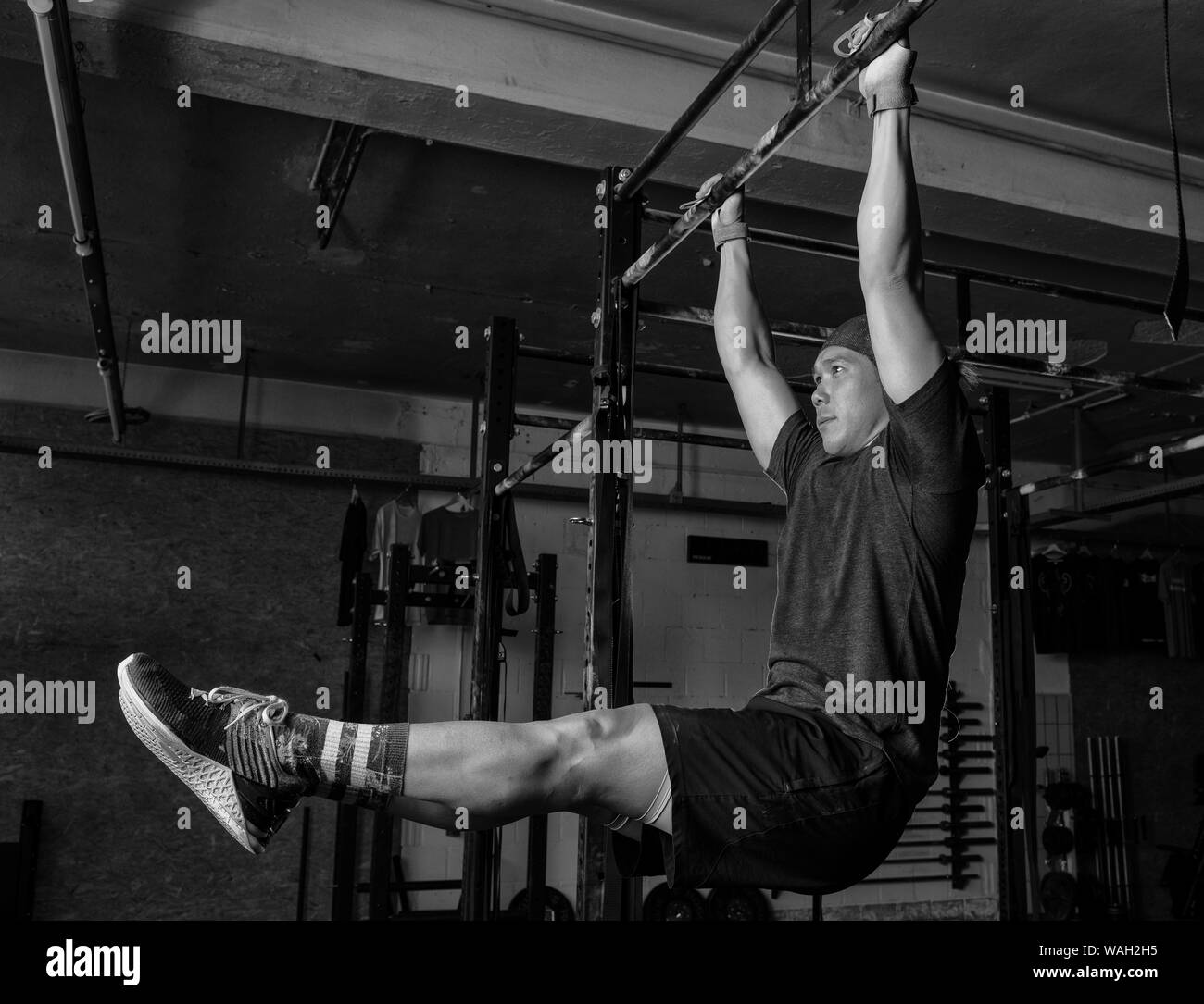 An attractive young man with muscles is doing the exercise l sit on the horizontal bar. Functional fitness workout in a gym. Black and white. Stock Photo