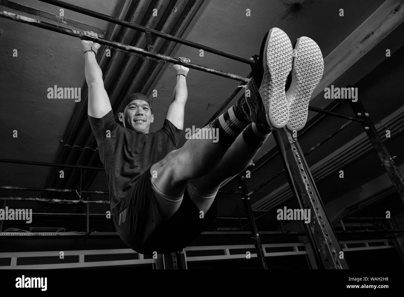 An attractive young man with muscles is smiling and doing the exercise l sit on the horizontal bar. Functional fitness workout in a gym. Stock Photo