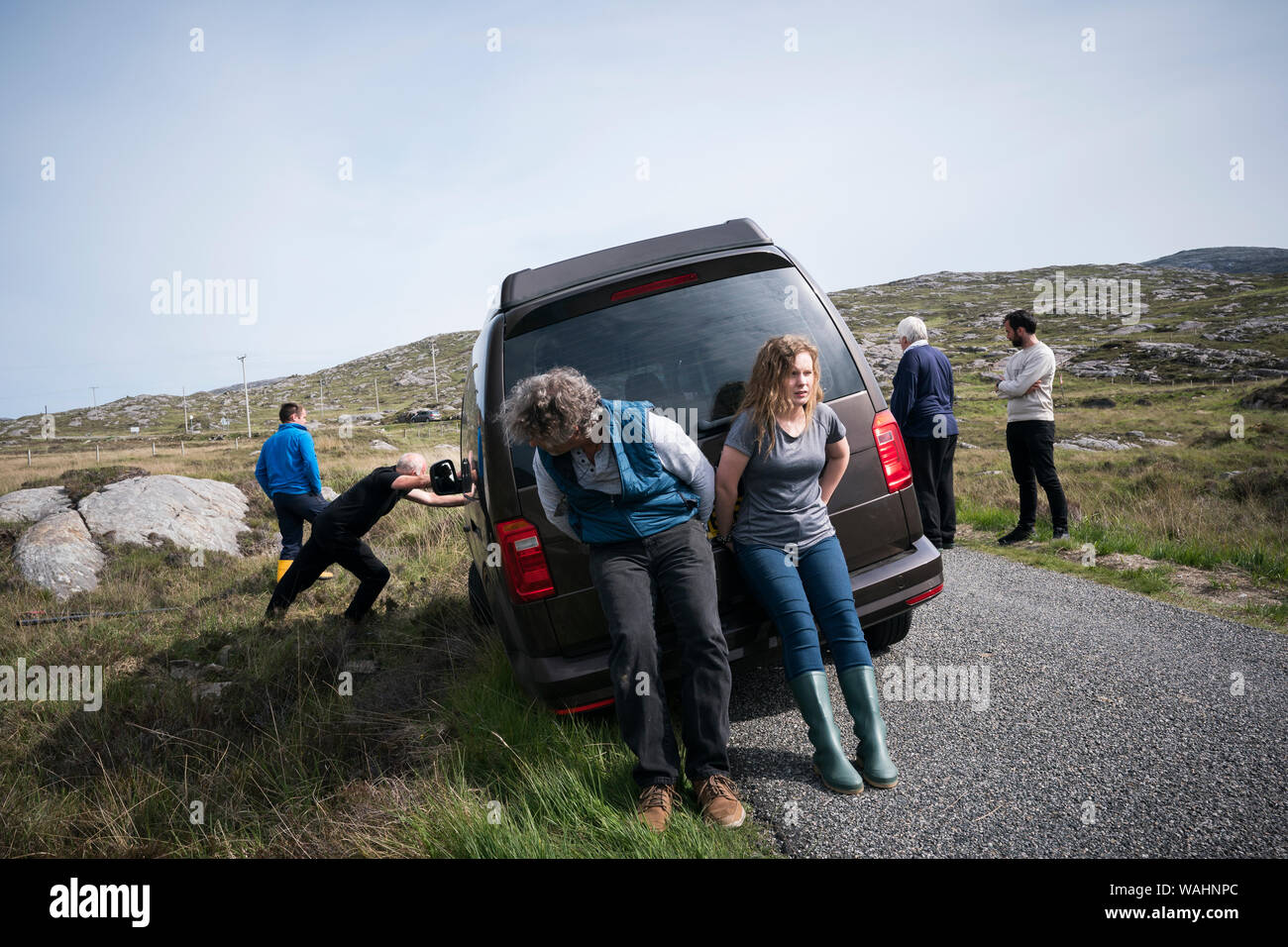 Man and teenage girl push the back end of a car rental stuck in the ditch as other tourists watch from the sidelines as another adult man pushes the f Stock Photo