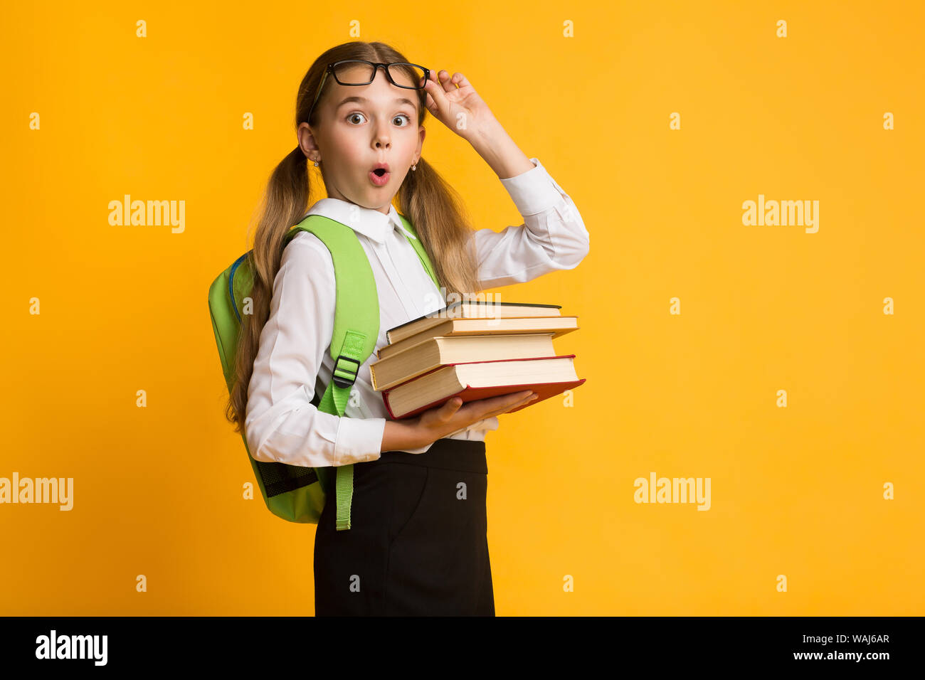Shocked Primary School Girl Holding Stack Of Books, Yellow Background Stock Photo