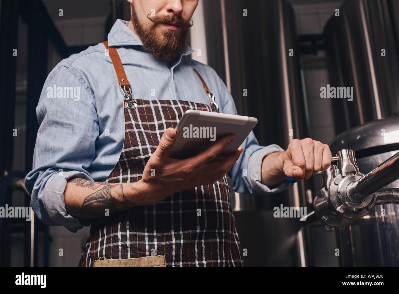 Man changes technological settings at the brewery according to the standarts listed at the tablet screen. Stock Photo