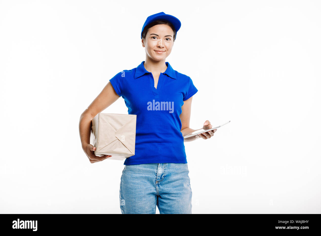 Postman in the blue uniform poses at the camera with a parcel and a tablet on the white background. Stock Photo
