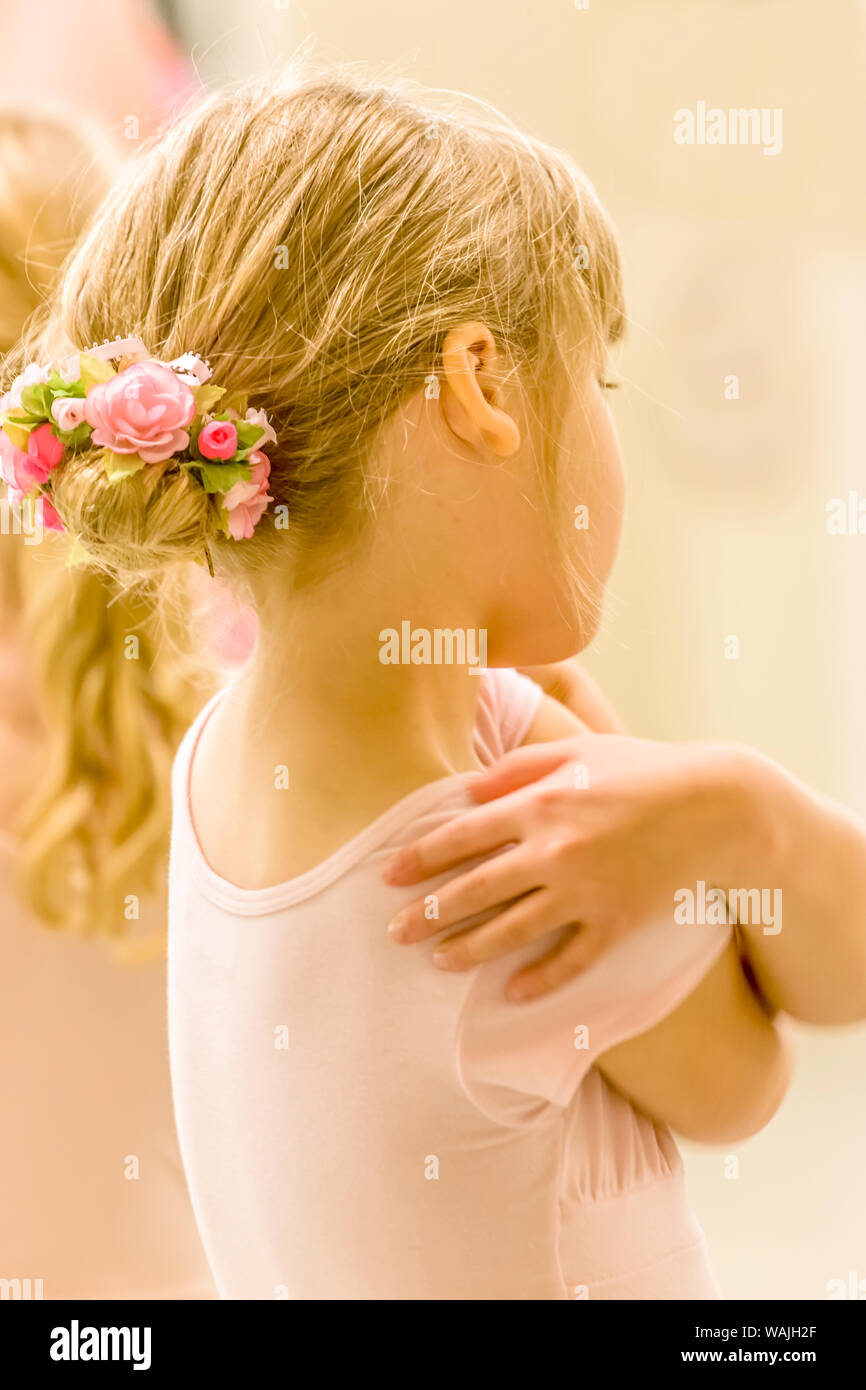 Rear view portrait of a six year old girl ballet dancer hugging herself, who is wearing a floral ballet bun hairpiece, in a dance studio. (MR) Stock Photo