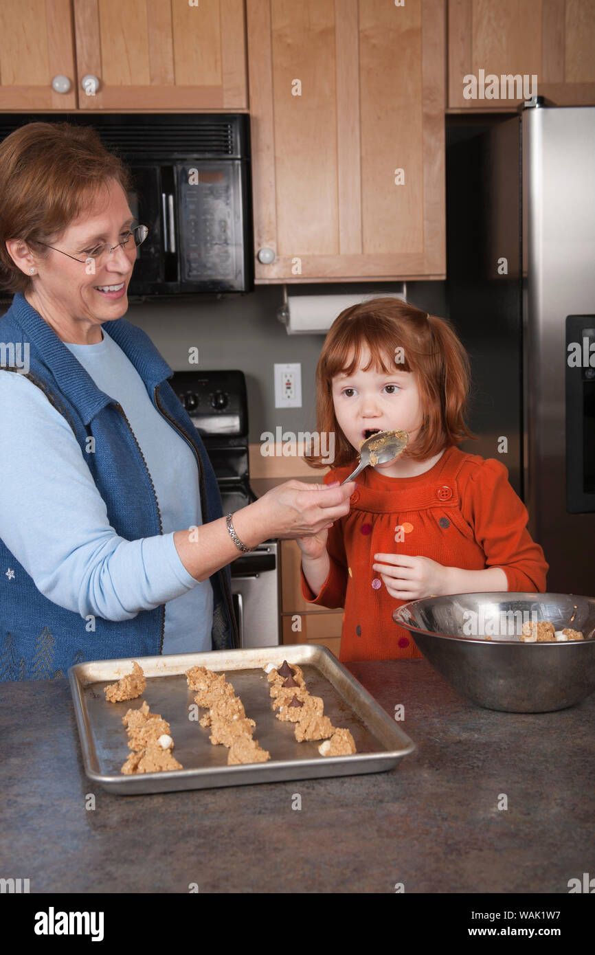 Grandmother holding spoon for 4 year old granddaughter to lick, while making cookies. (MR, PR) Stock Photo