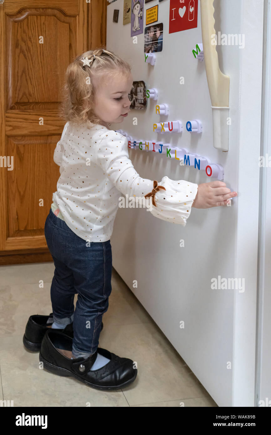 2 year old wearing adult shoes, at play with magnetic alphabet letters on her refrigerator. (MR,PR) Stock Photo