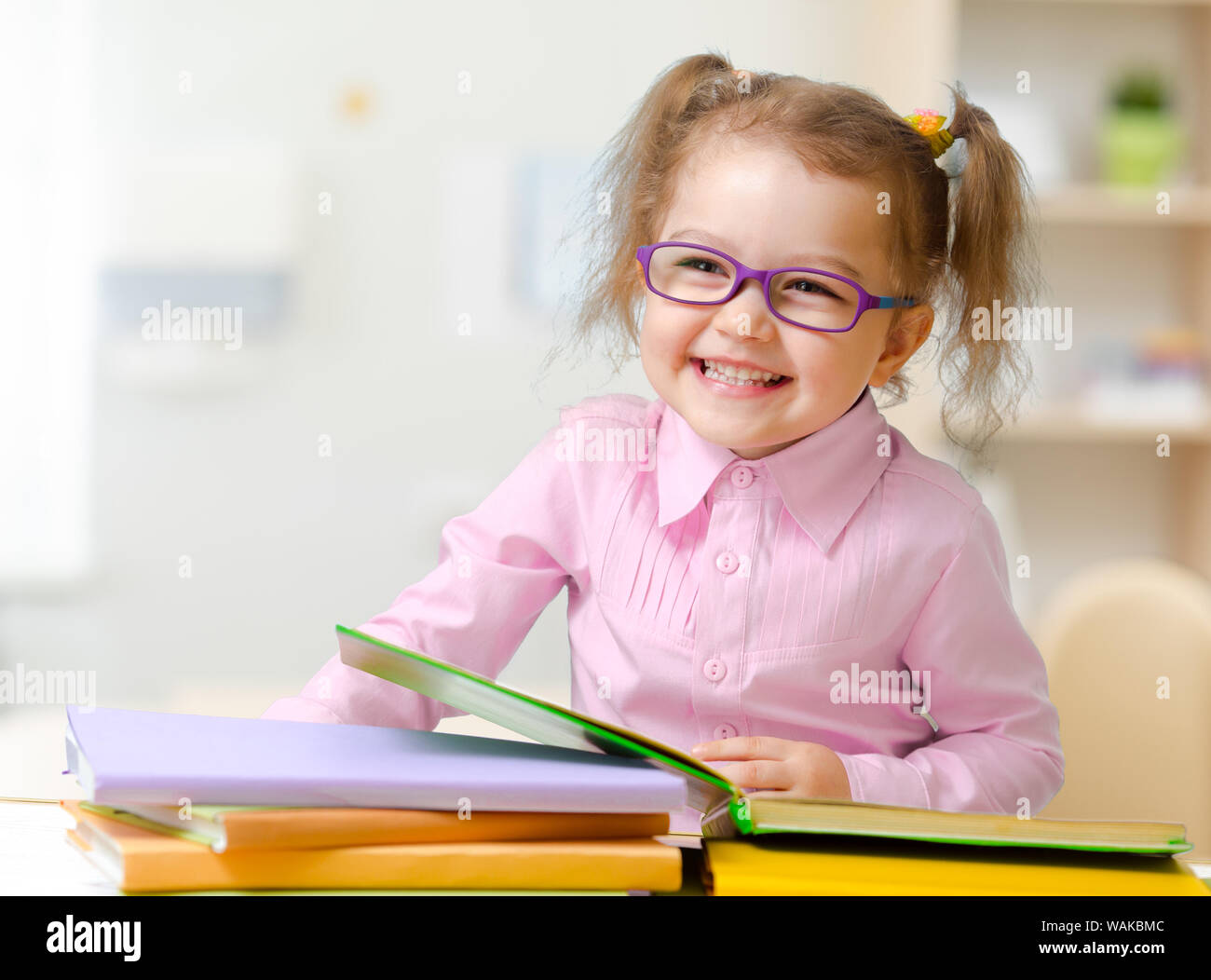 Happy kid girl in spectacles reading books sitting at table at her room Stock Photo