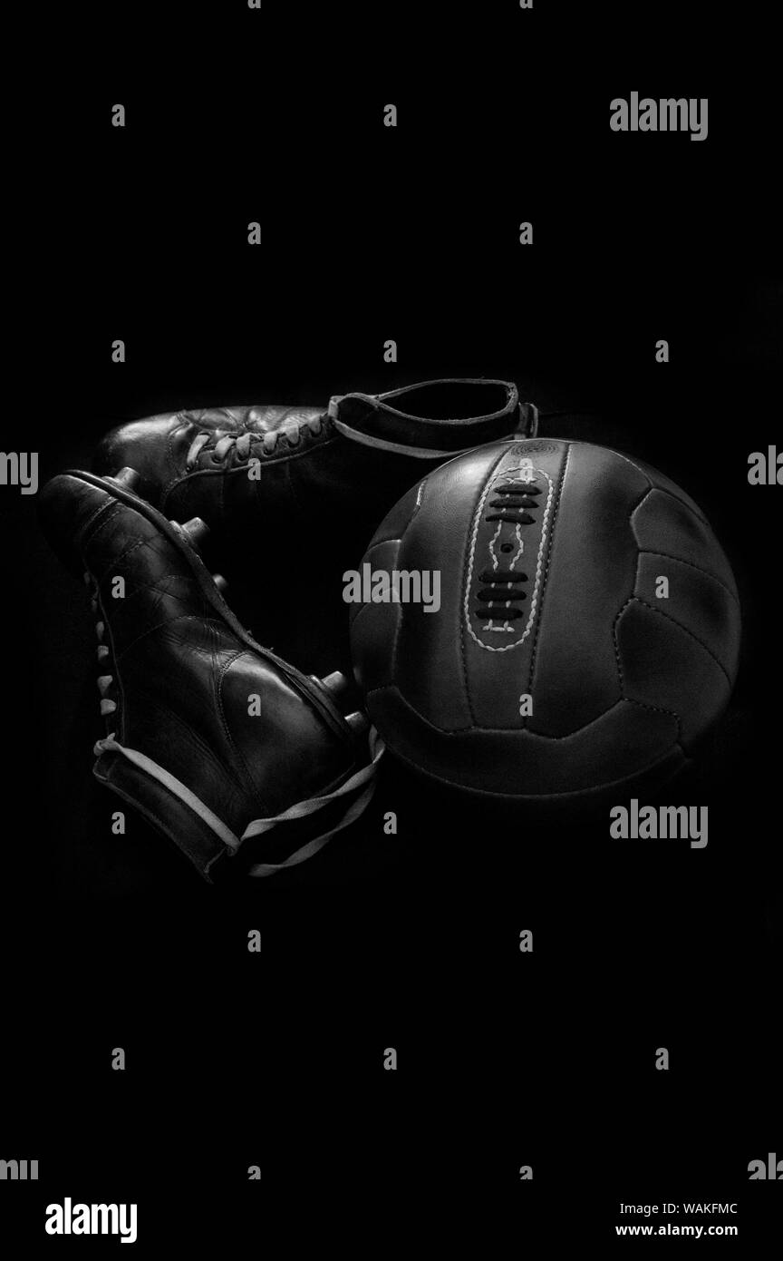 Antique leather football soccer ball and vintage cleats, boots, football shoes against a black background in black and white B&W Stock Photo