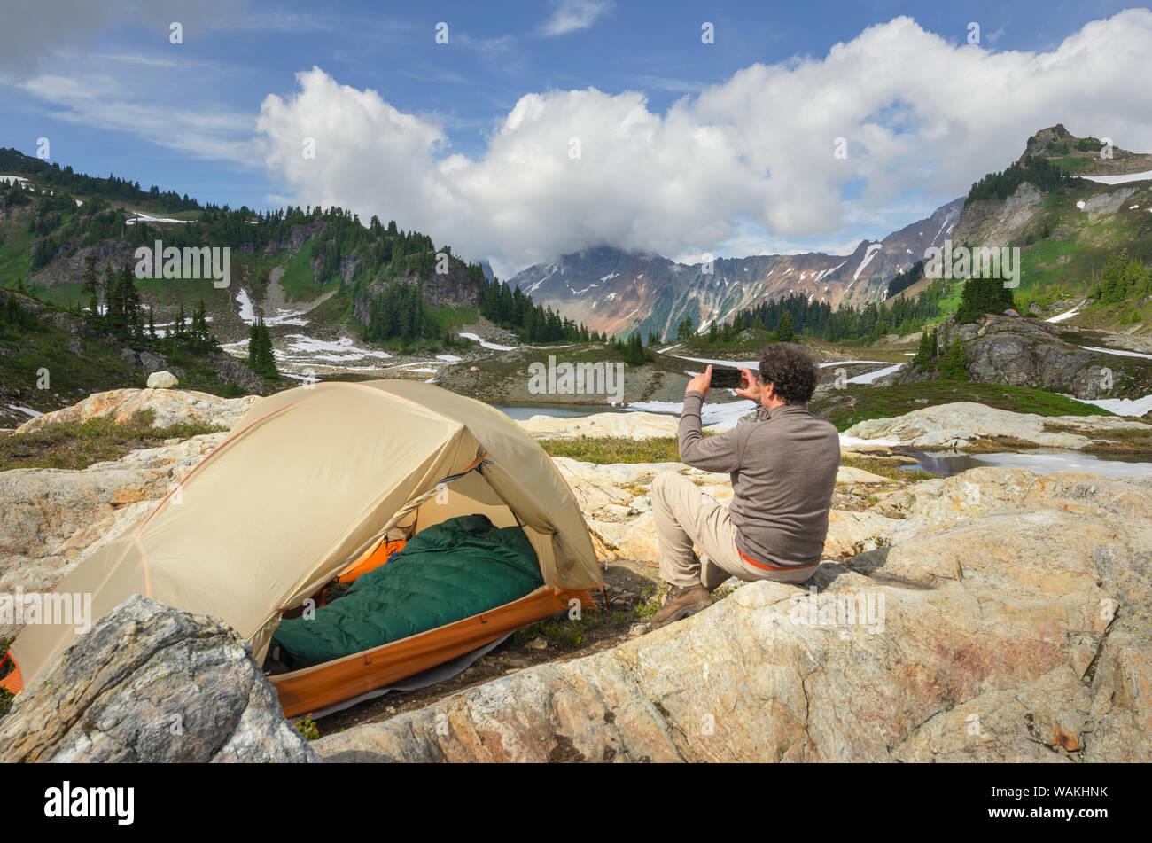 Man taking pictures with smartphone at backcountry campsite. Yellow Aster Butte Basin, Mount Baker Wilderness. Mount Shuksan is in the distance. North Cascades, Washington State Stock Photo