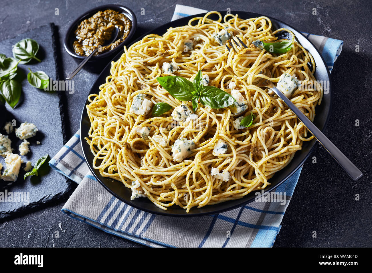 close-up of delicious Spaghetti with basil pesto and blue cheese on a black plate on a concrete table with pesto in a black bowl, Italian cuisine, hor Stock Photo