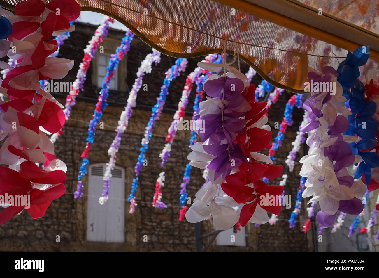 A partial view of a traditional stone French house through flower festival decorations hanging from a shop awning Stock Photo
