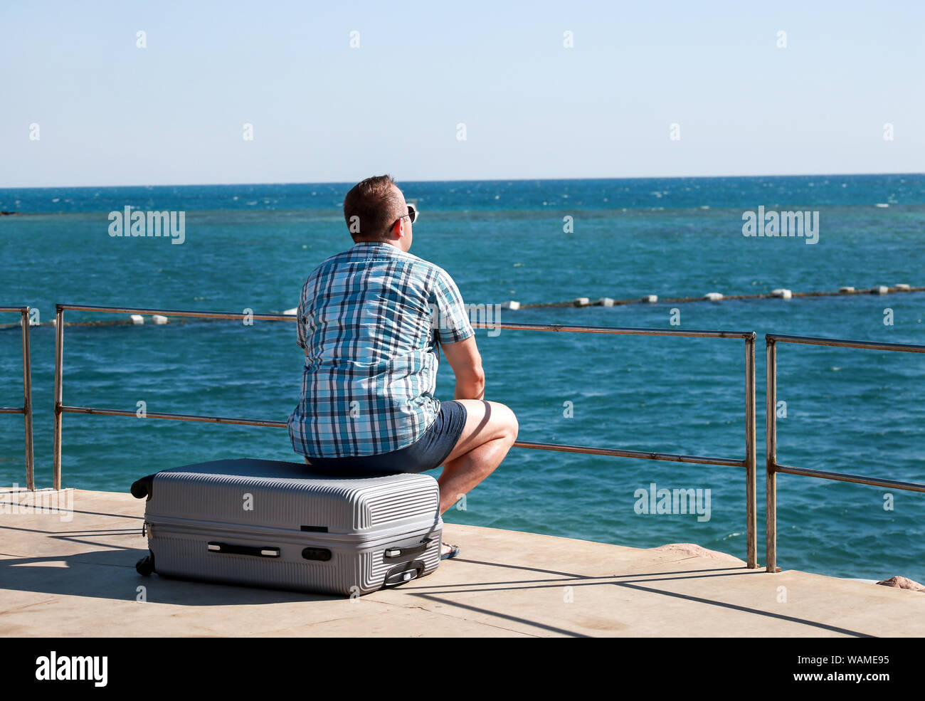 Tourist and traveler is sitting on his traveled suitcase on beach of hotel resort, enjoys view sea environment, nature landscape, panorama, horizon. Stock Photo