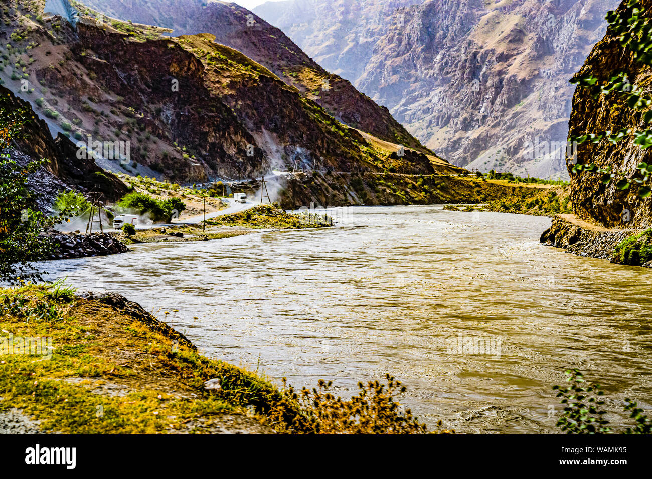 Trucks from China travel along the new Silk Road beside the Panj River near Toghmay, Tajikistan, directly on the Afghan border. On the opposite bank in Afghanistan, only pack animals and smaller vehicles are seen on the road. Stock Photo