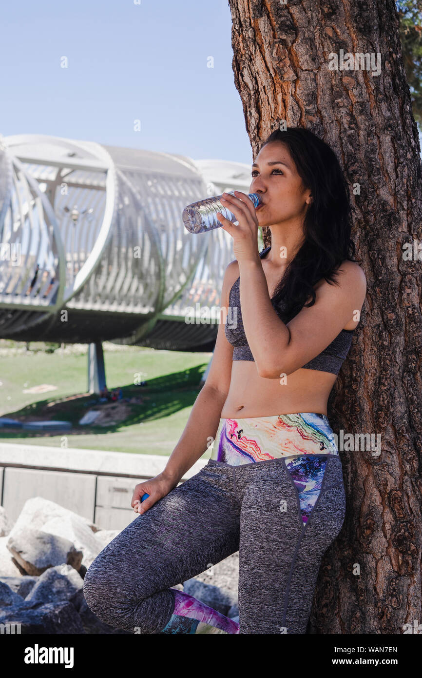 A young woman stays hydrated by taking a drink of water from a bottle in the middle of a workout in the park in front of a picturesque bridge Stock Photo
