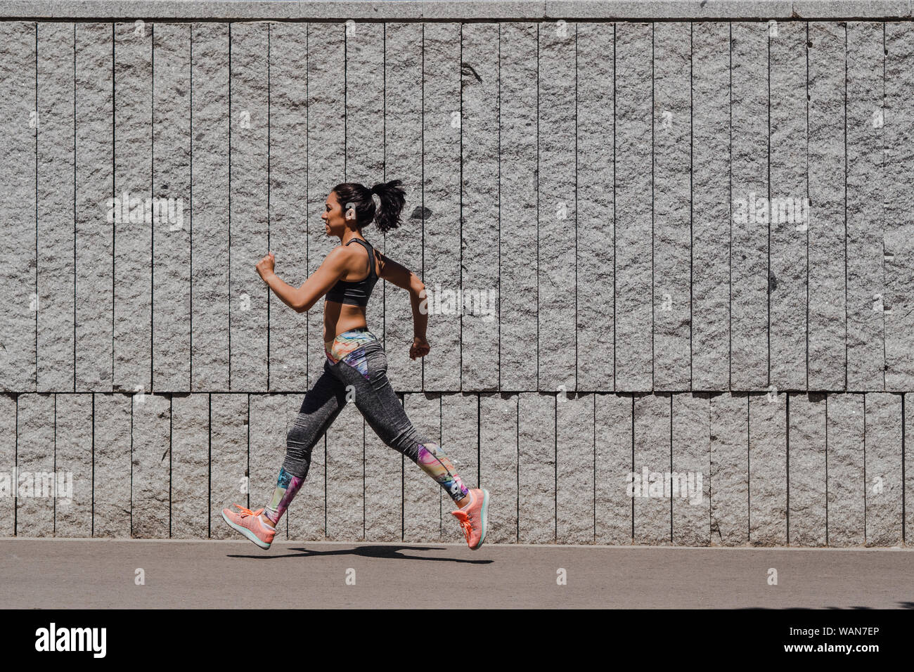 Side view of a woman dressed in sportswear running fast in front of a gray wall Stock Photo