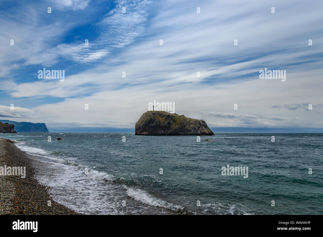 Rock with a cross stands in the sea against the cloudy sky Stock Photo