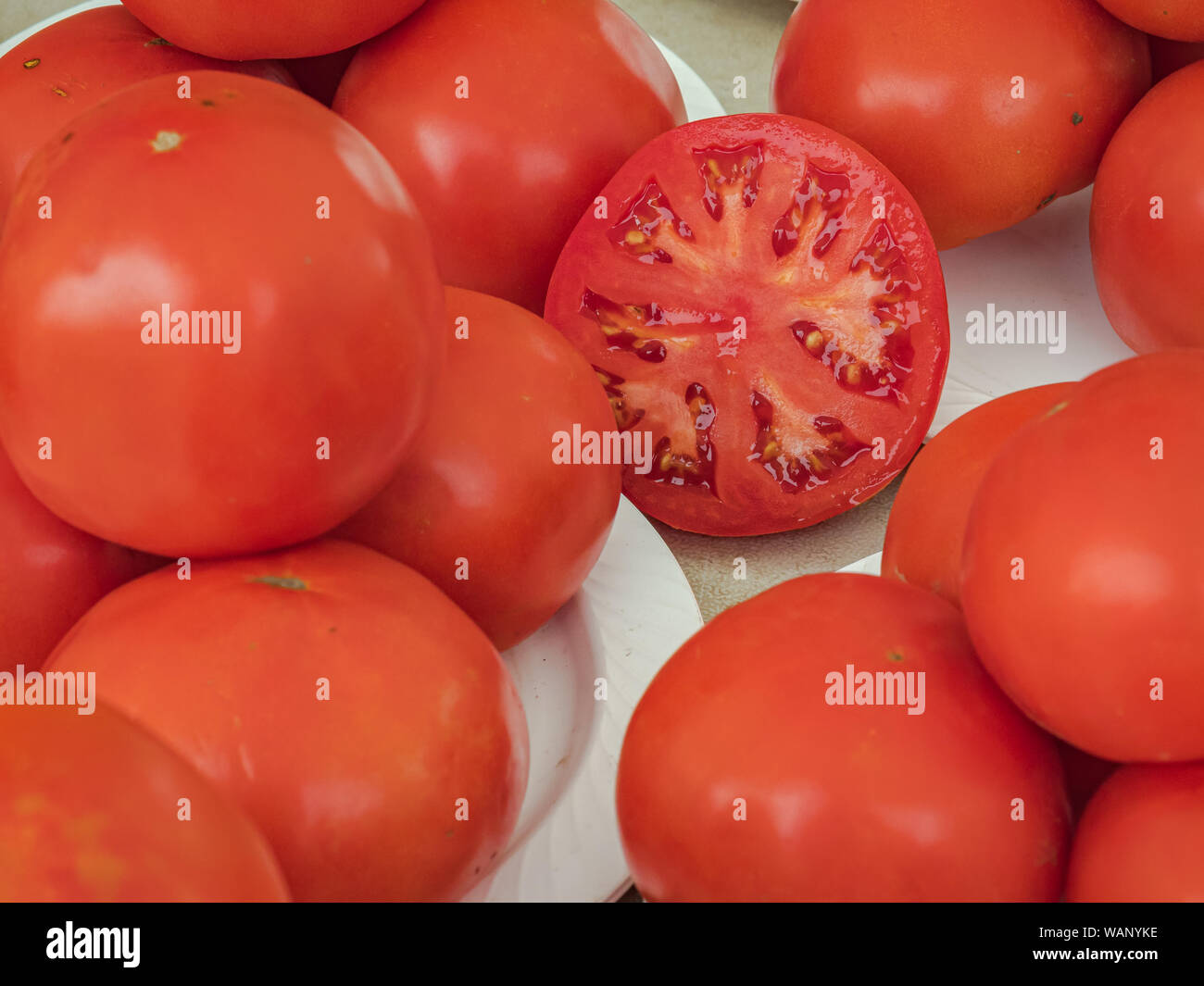 One sliced tomato on a white table with piles of  whole ripe red tomatoes. Stock Photo
