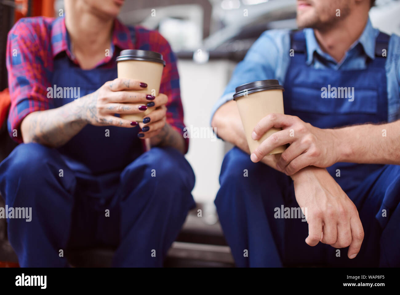 Close up of two automechanics in tartan shirts drinking coffee from paper cups during their lunch break. Stock Photo
