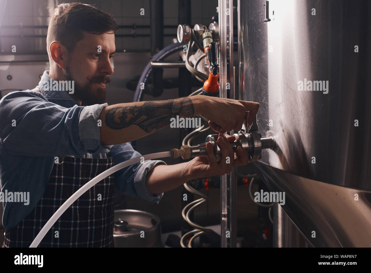 Brewery worker puts on a pipe onto the tap of a big chrome barrel in the dark plant room. Stock Photo