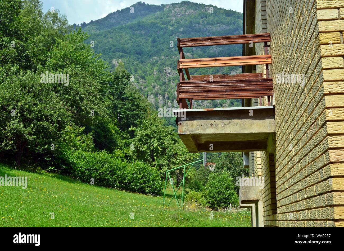 A wooden balcony of a yellow brick mountain village house with a basketball outdoor court and a mountain in the background, a forest on the left side Stock Photo