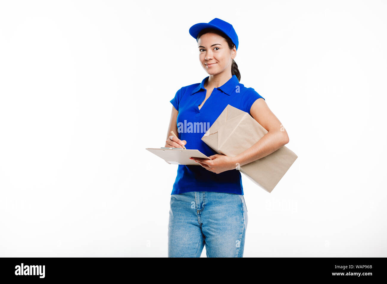 Girl working in the delivery wears blue uniform as she signes documents for the parcel she holds. Stock Photo