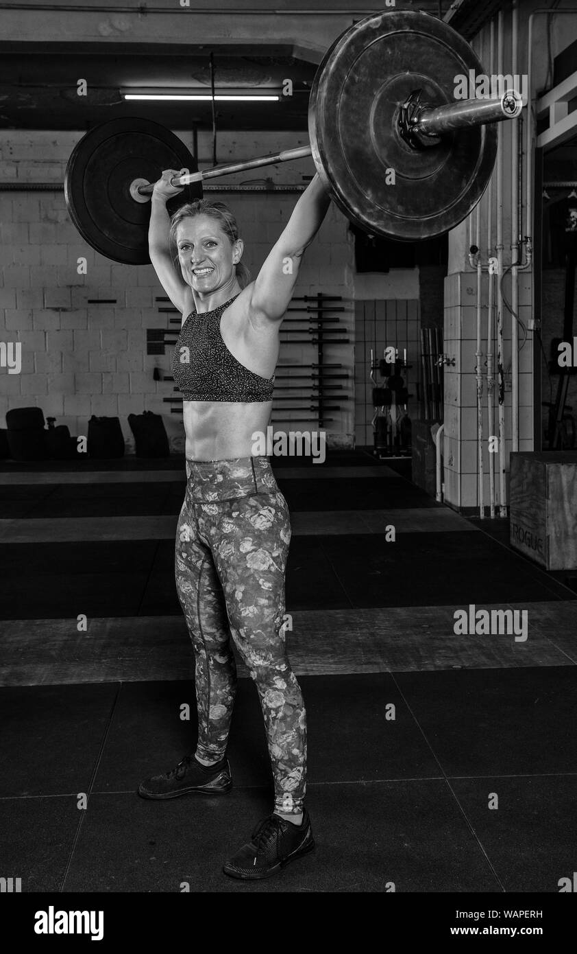 An handsome middle aged blonde woman with Sixpack is doing an overhead clean with the barbell. Functional fitness and weight lifting workout in a gym. Stock Photo