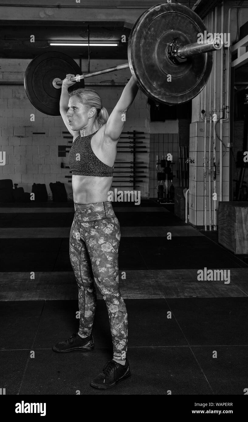 An handsome middle aged blonde woman with Sixpack is doing an overhead clean with the barbell. Functional fitness and weight lifting workout in a gym. Stock Photo