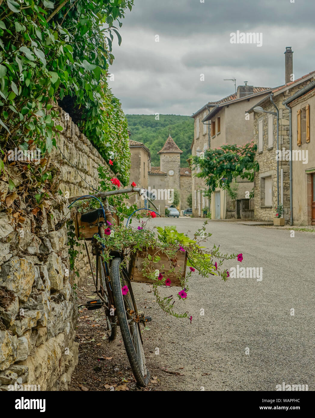 Feneyrols, Midi Pyrenees, France - July 23, 2017: Front view of an old bicycle decorated with flowers with a bell tower at the bottom of the street Stock Photo