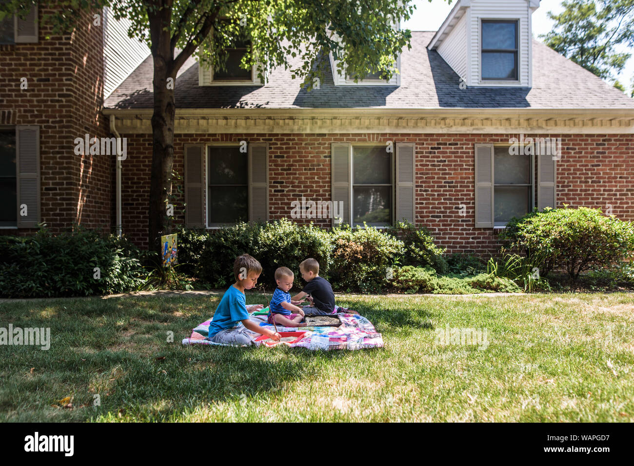 3 boys on a quilt in front yard of brick house doing school work Stock Photo