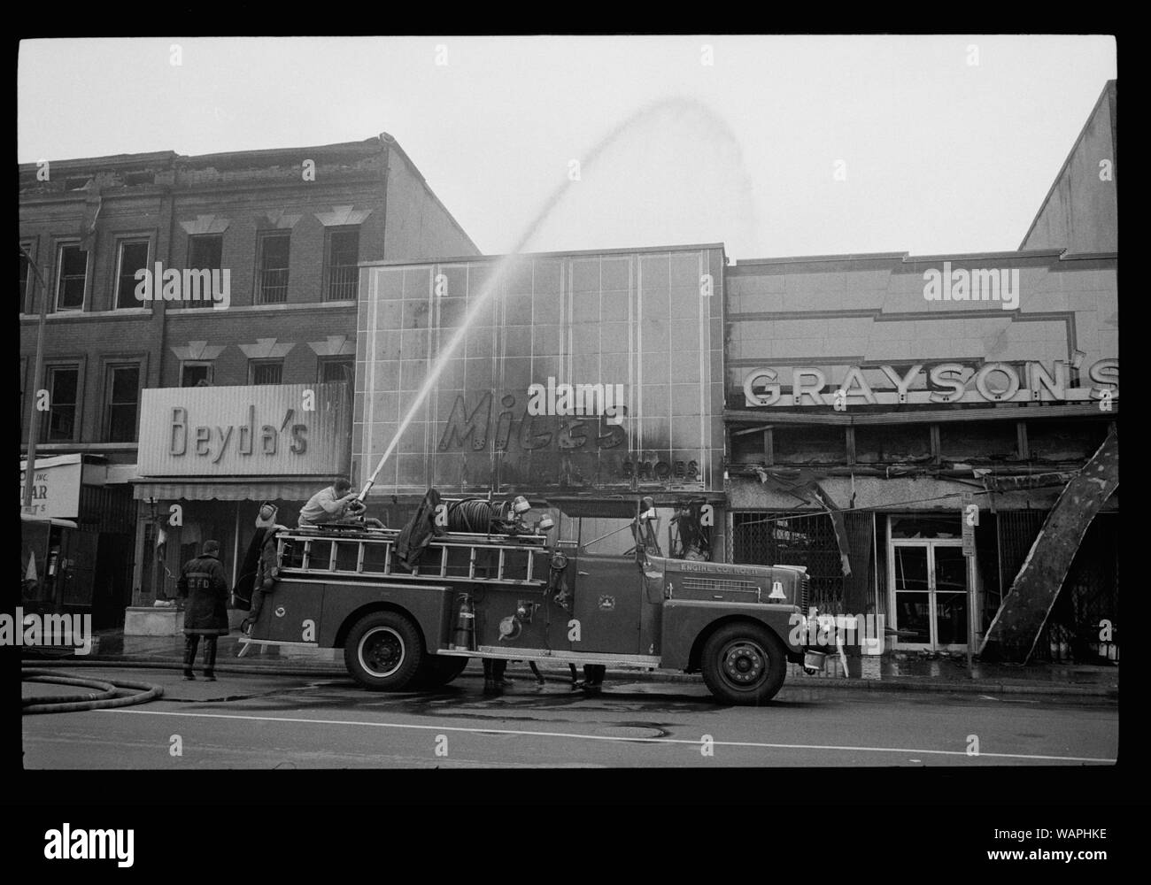 D.C. riot. April '68. Aftermath; English: Photograph showing firefighters spraying water on shops, including Beyda's, Miles Shoes, and Graysons, that were burned during the riots that followed the assassination of Martin Luther King, Jr. Stock Photo
