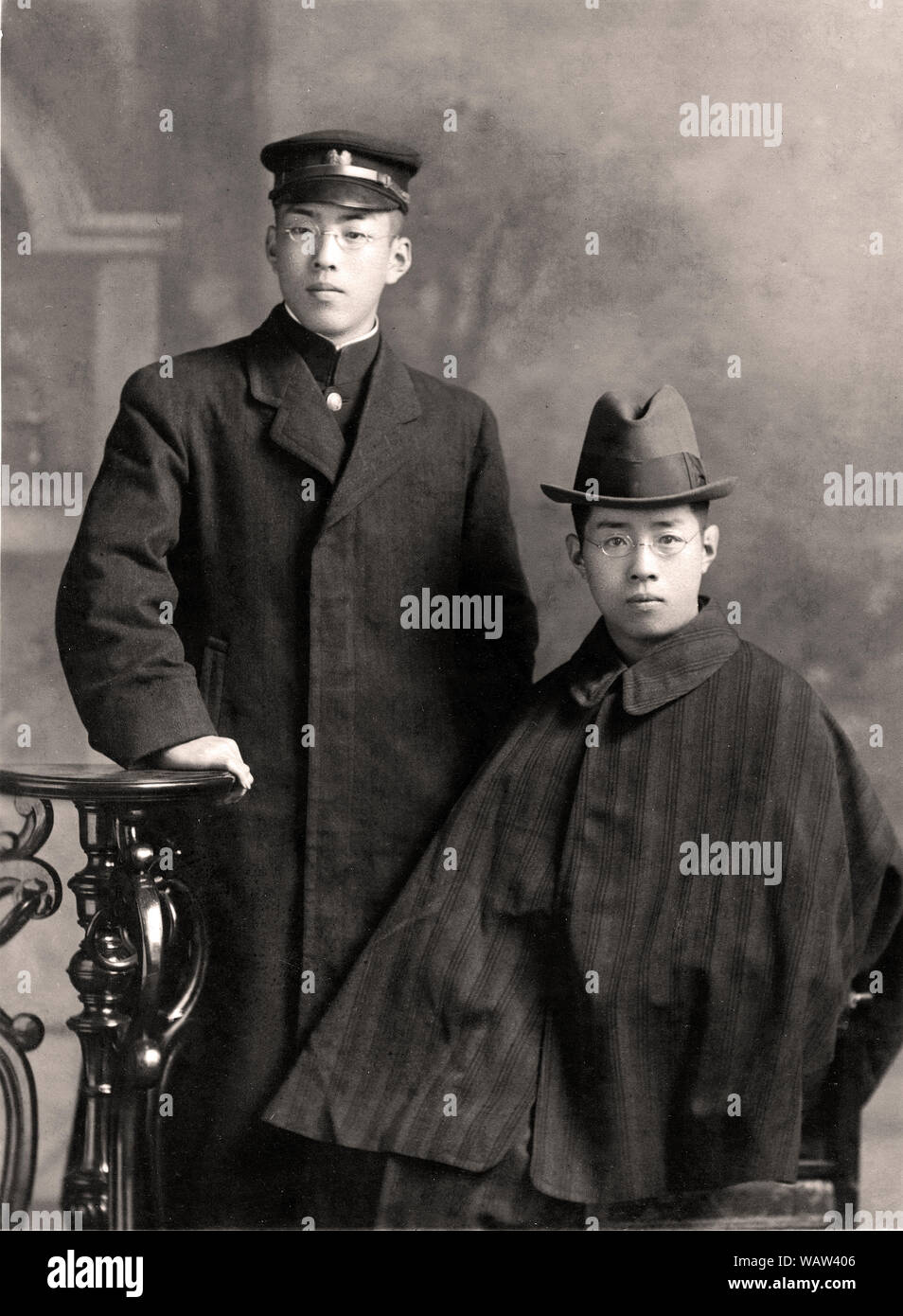 [ 1910s Japan - Japanese Tonbi Coat and Uniform ] —   Two brothers, 20 and 21 years old in December 1912 (Taisho 1). One wears a university uniform, the other an Inverness style tonbi coat.   The tonbi coat, a perfect fusion of Japanese and Western styles, was first introduced around 1887 (Meiji 20), but reached its zenith of popularity during Taisho (1912-1926) and early Showa (1926-1989). The coat was especially popular with intellectuals.  20th century vintage gelatin silver print. Stock Photo