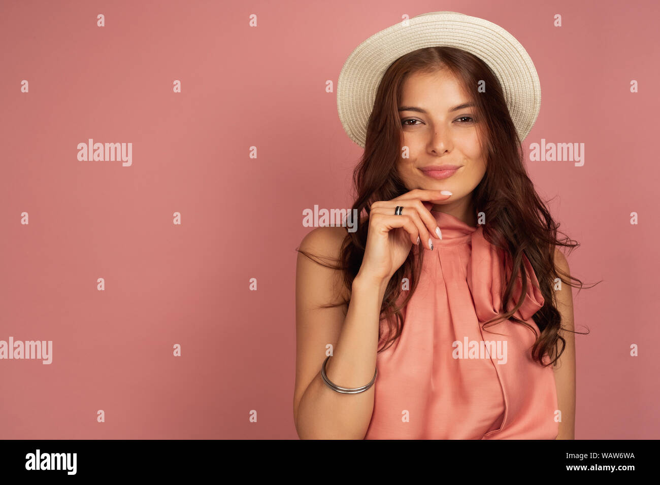 A dark-haired girl in a pink dress, and a straw hat on a pink background, propping her chin with her eyes squinting at the camera. Stock Photo