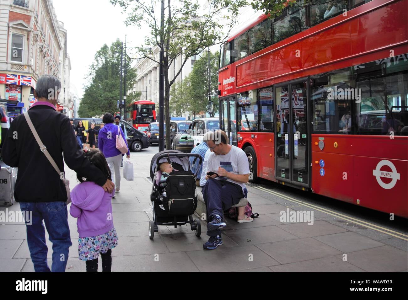A man is sitting on a bench in Oxford Street on a busy day while looking after a baby in a pram, London, UK Stock Photo