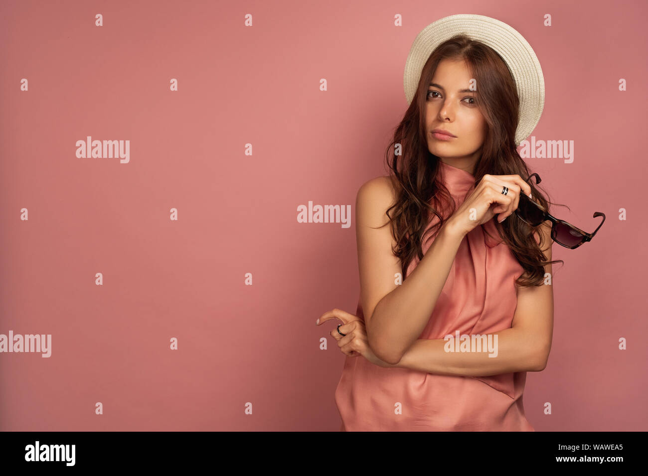 A dark-haired girl in a pink dress and a straw hat holds in glasses and looks into the frame, her head bowed to the side. Stock Photo