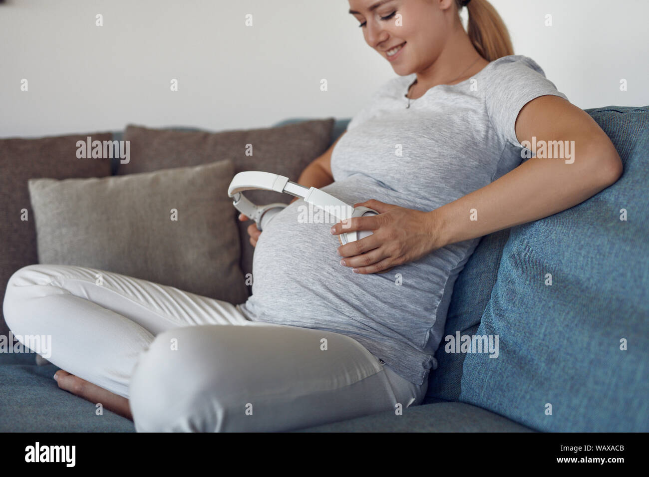 Smiling happy pregnant young woman providing music with earphones to her unborn baby by holding a set of stereo headphones to her abdomen as she sits Stock Photo