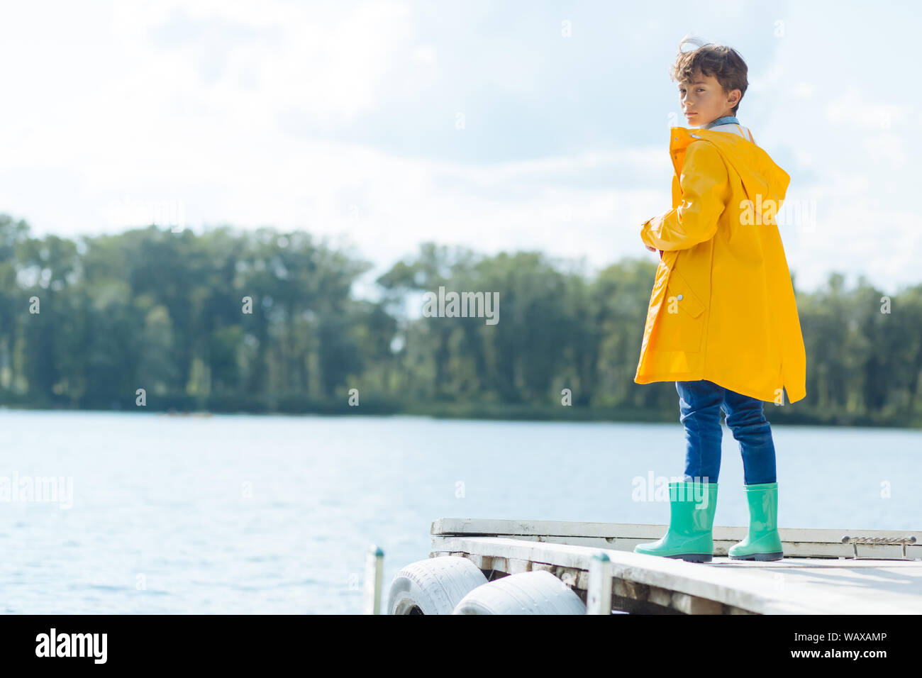 Dark-haired boy wearing raincoat standing near river Stock Photo
