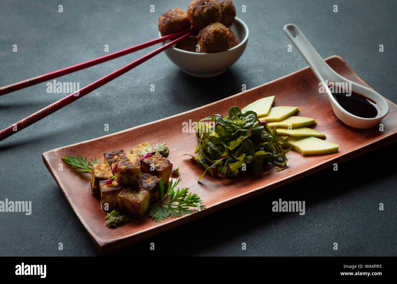 fried tofu on wooden plate with vegetables, sauce and avocado with falafel and chopstick on the dark background Stock Photo