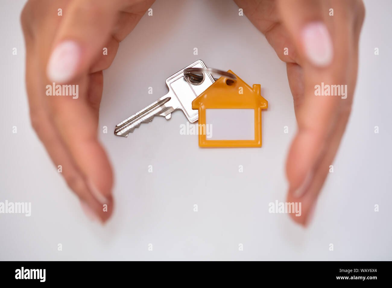 Close-up Of Person's Hand Protecting House Key With Keychain On White Desk Stock Photo