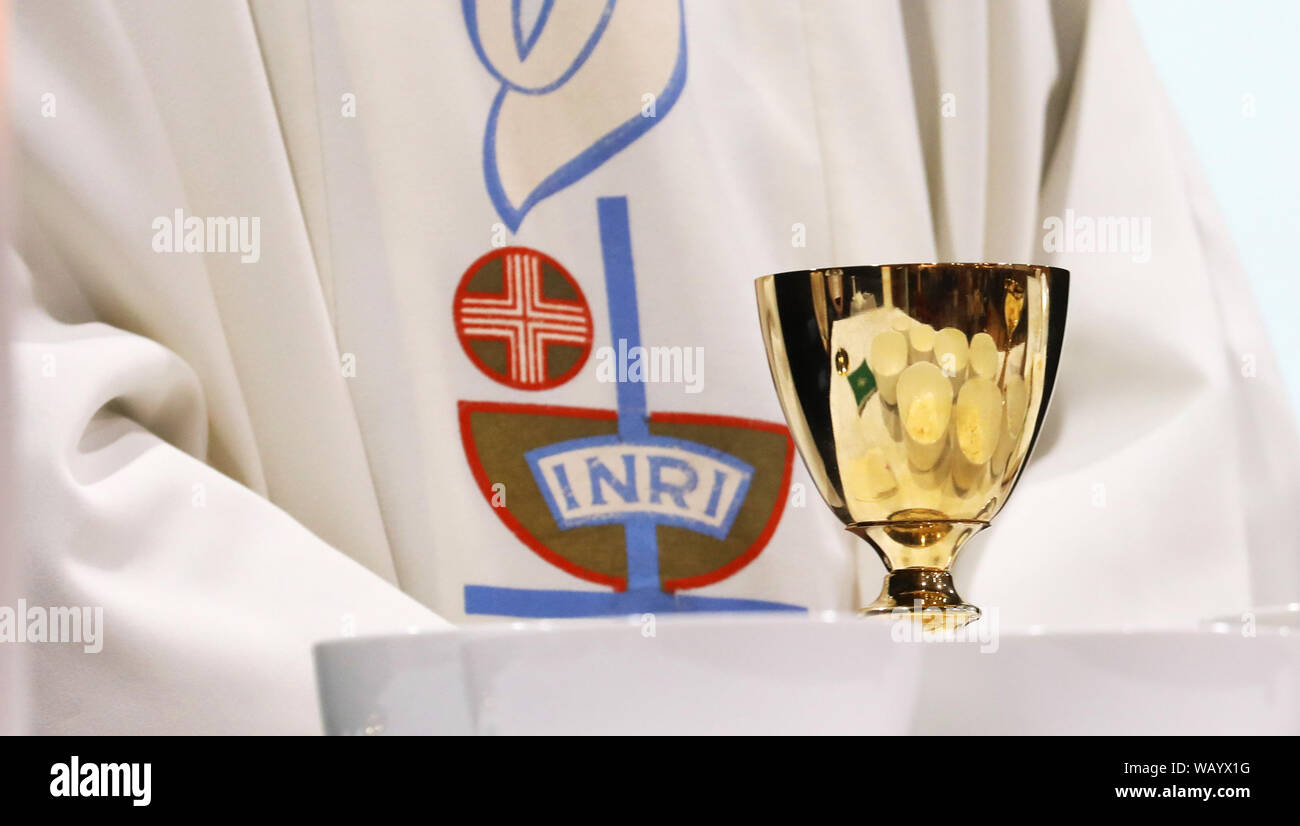 Close up of a priest wearing a white gown with the word INRI embroidered on to it. A single brass chalice with reflected bowls containing the host Stock Photo