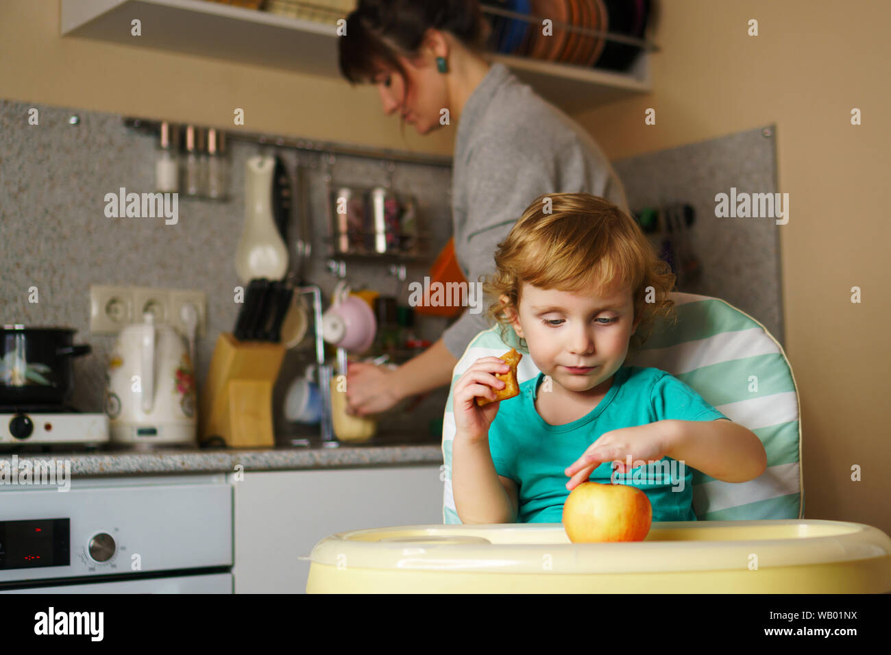 Adorable little baby eating dessert in the kitchen. He sits in a highchair. Mom in the background defocusing the dishes. Weekday young family. Materni Stock Photo