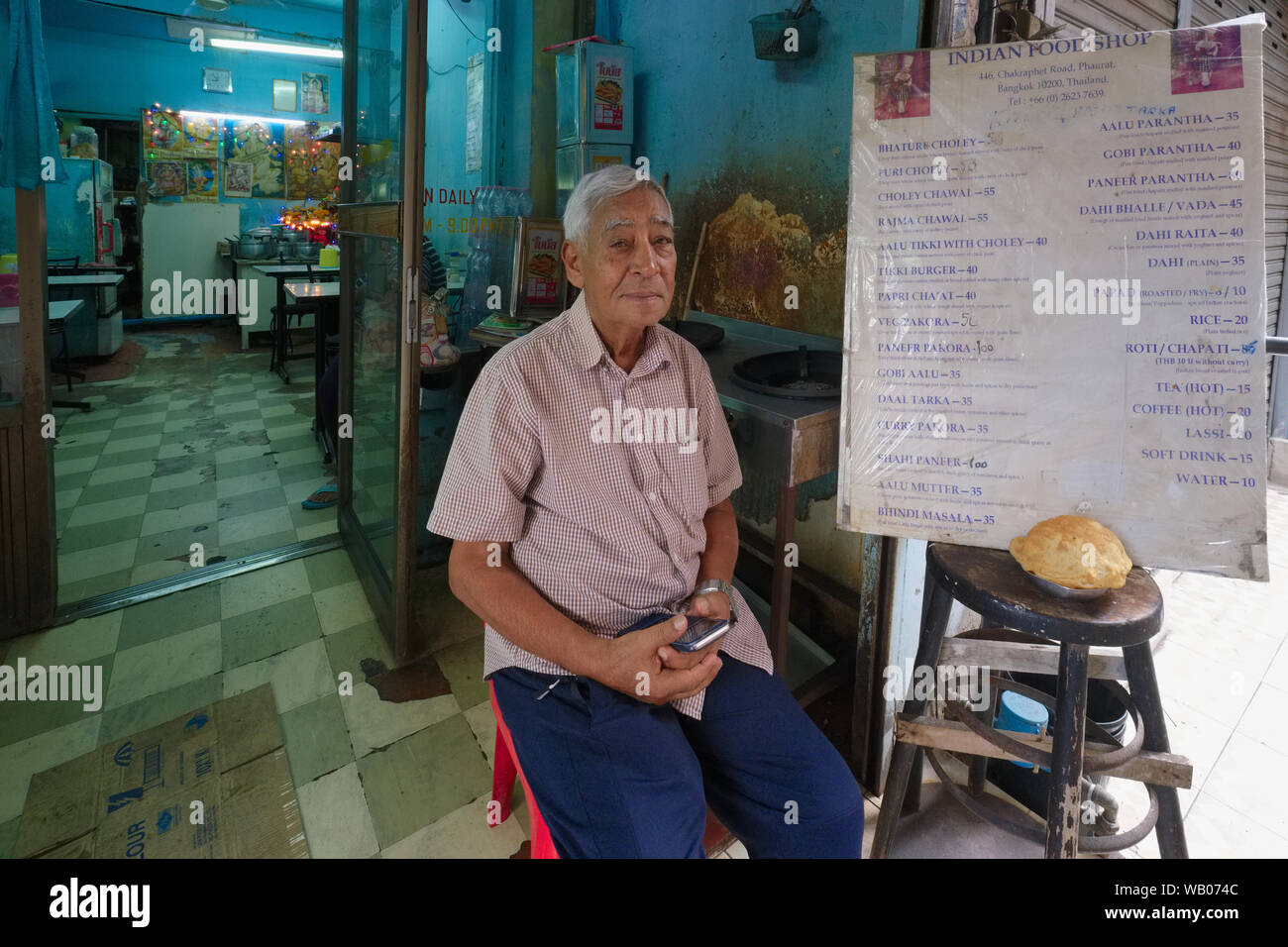 The Indian proprietor sitting outside his small Indian eatery at Pahurat or 'Little India', located next to Chinatown, Bangkok, Thailand Stock Photo
