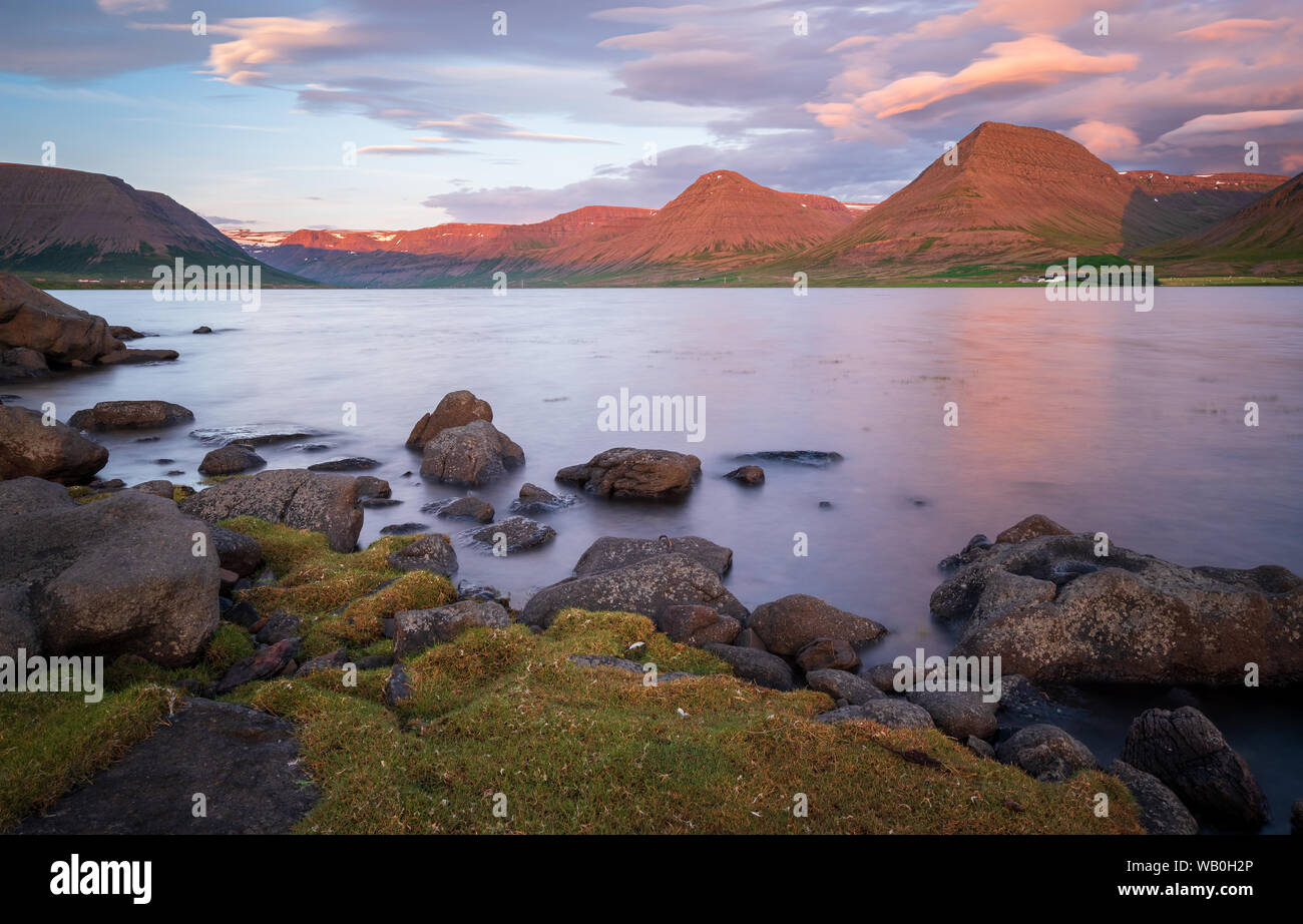 Summer sunset at coast of Westfjords with rocks and green banks in foreground and beautiful lighted mountains in background, Vestfirðir, Iceland Stock Photo