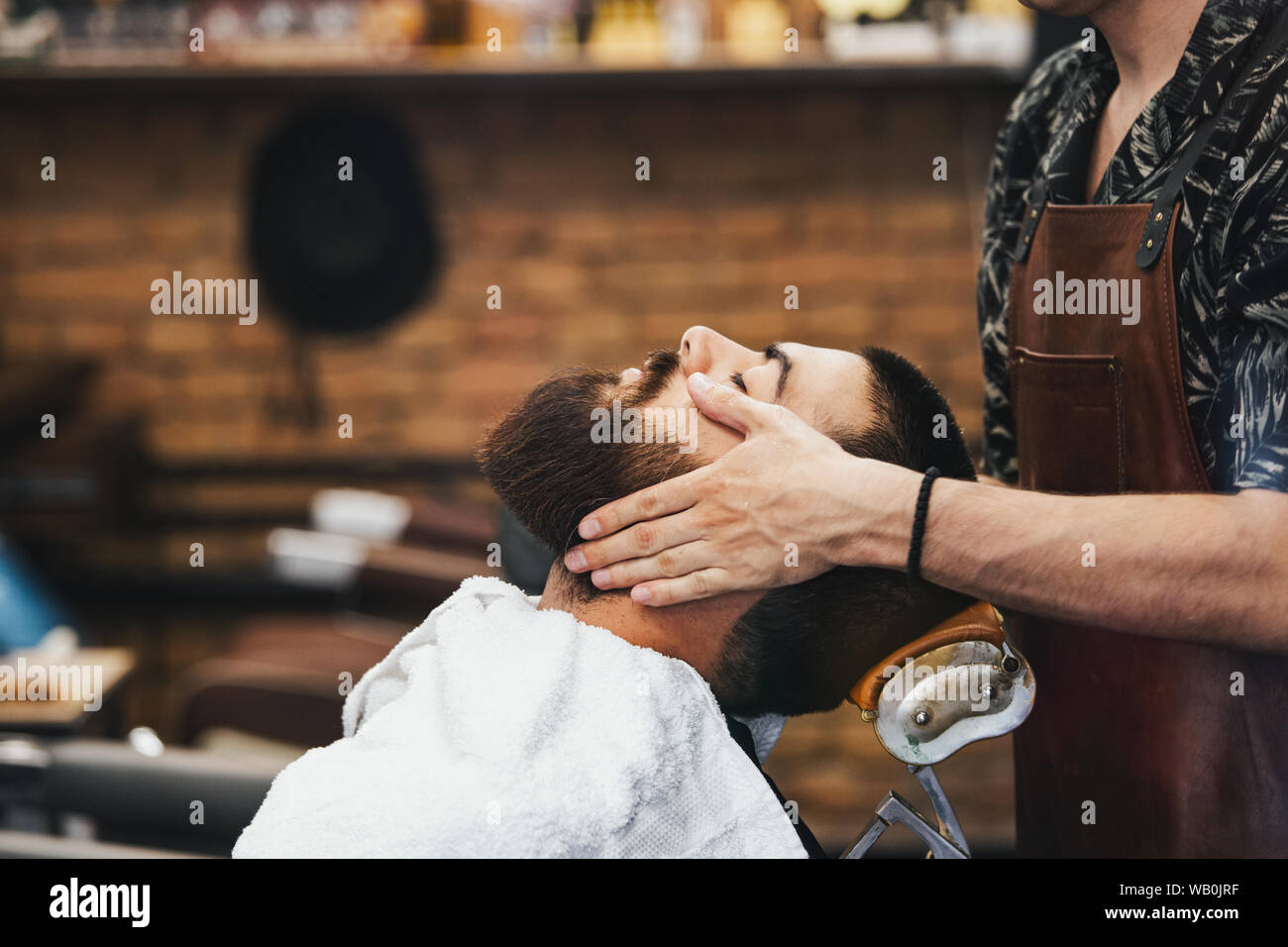 Handsome man with a beard and closed eyes in a black cutting hair cape in the barbershop. Barber in is doing him a face massage. Closeup. Horizontal. Stock Photo