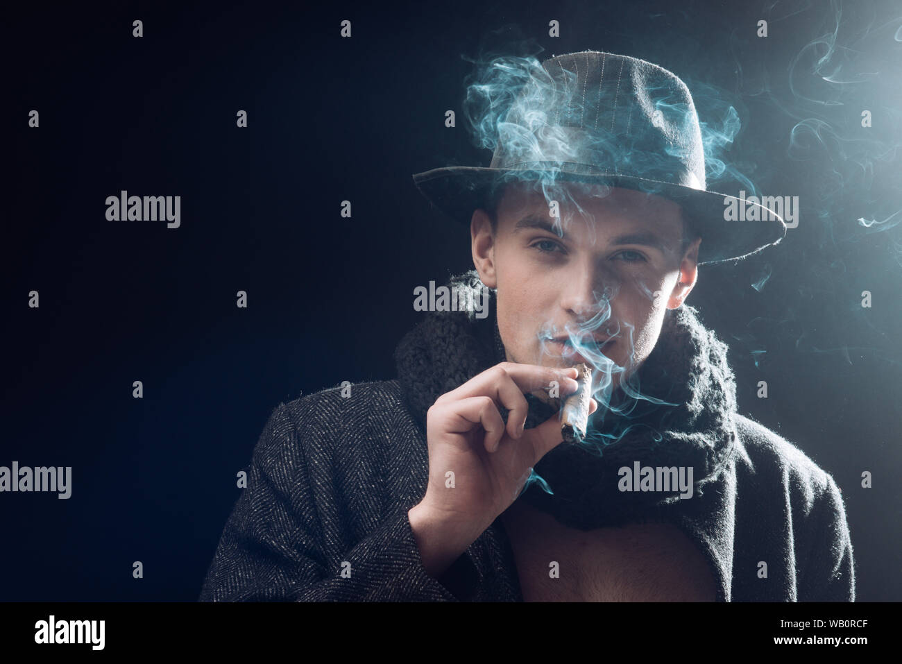 Man in coat, hat smoking cigar, dark background. Macho on mysterious face, detective, investigator, agent. Guy in old fashioned outfit looks mysteriou Stock Photo